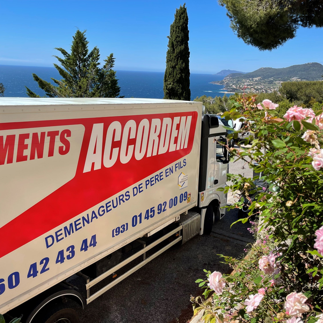 Camion de déménagement Accordem stationné avec vue sur la mer Méditerranée et les collines, entouré de fleurs roses en été