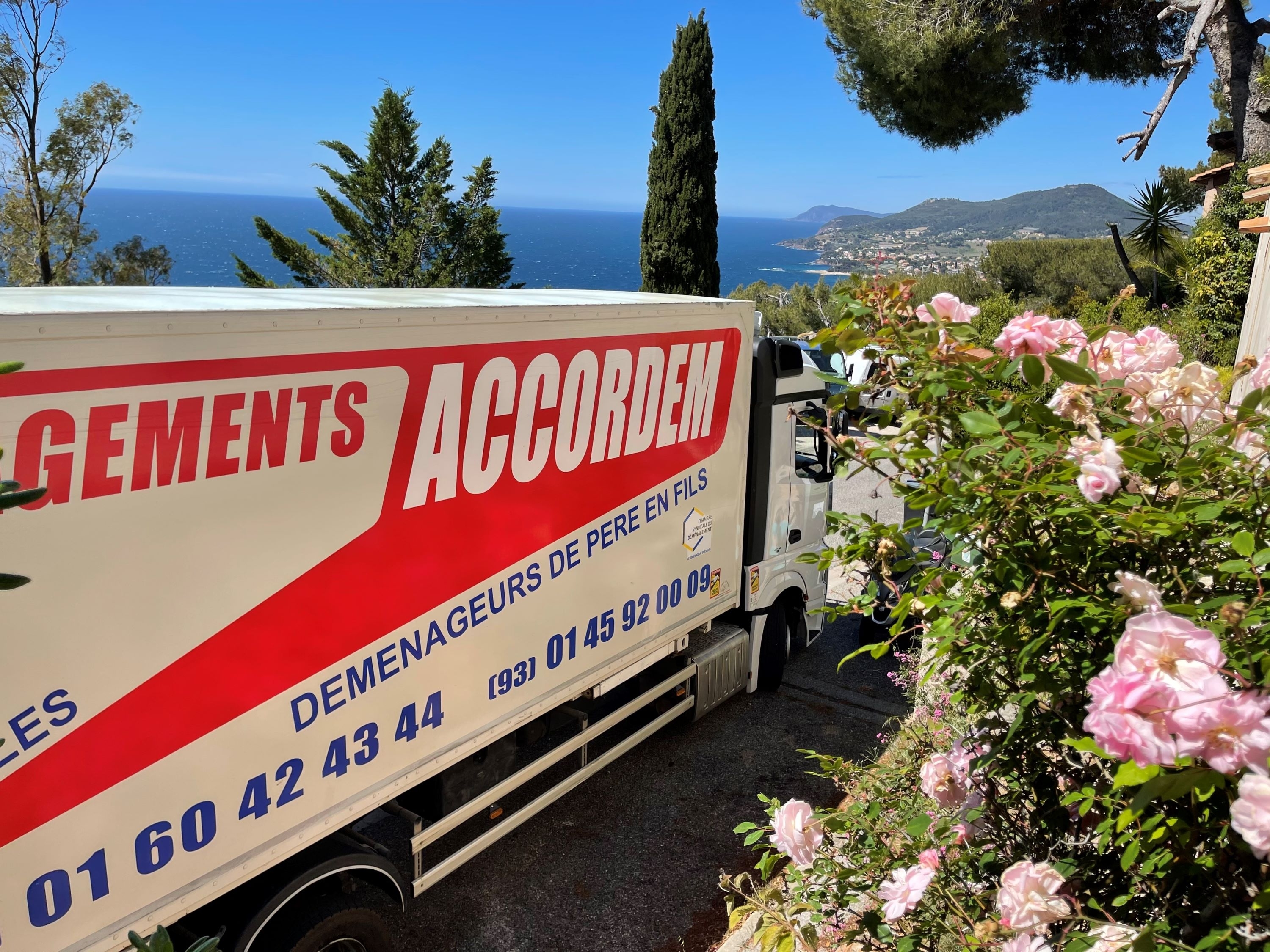 Camion de déménagement Accordem stationné avec vue sur la mer Méditerranée et les collines, entouré de fleurs roses en été